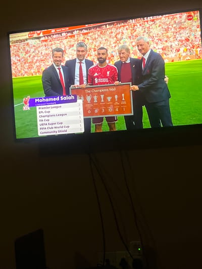 The image shows a television screen displaying a graphic about Mohamed Salah's achievements with Liverpool FC. The graphic lists his honors, including Premier League titles, FA Cups, Champions League titles, and others. Salah is pictured in his Liverpool kit. People in suits are also shown holding up a Champions Wall with trophies listed. The background is a soccer stadium with fans. The room is dimly lit and the image is taken at an angle. There is a white power outlet with several cords coming from it visible in the right. No location in Oshogbo, Nigeria is evident in the image.