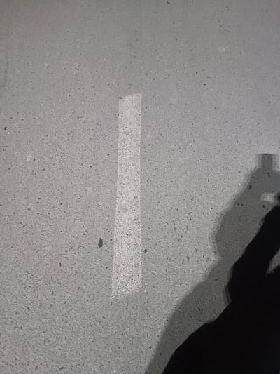 A gray, textured surface is bisected by a vertical, rectangular highlight. The surface appears to be concrete or stone with small, speckled variations. A dark, blurred shadow of a hand extends from the bottom right corner, suggesting the image was captured indoors under artificial lighting. The light source is positioned in front, creating a defined shadow.