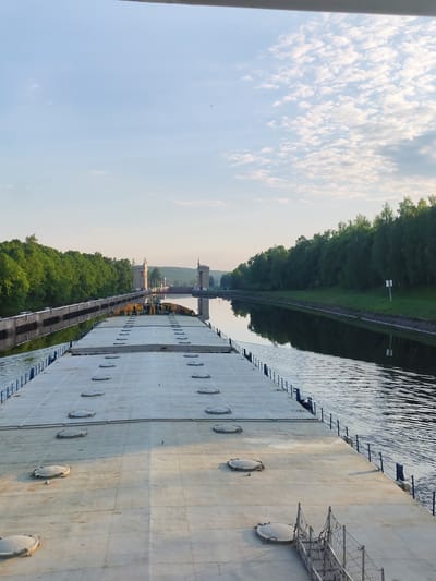 The image depicts a large, flat-decked barge traversing a canal in what appears to be a natural setting. The location is likely within or near the city of 3-Y Uchastok, Russia.
The barge occupies the foreground and extends into the midground. Its deck is composed of a series of rectangular panels, punctuated by numerous circular access hatches. A railing runs along the edge of the barge, separating the deck from the canal.
The canal, flanked by dense green trees on both sides, stretches toward a structure in the distance that could be a lock or other canal control facility. The sky above is partially cloudy, with a blue expanse visible between the clouds. The water in the canal is relatively calm, with reflections of the sky and surrounding trees.