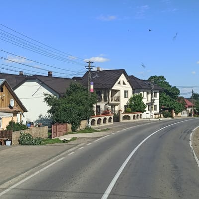 This is a street-level, outdoor, daytime photograph taken in Milișăuți, Romania. The image shows a two-lane asphalt road curving to the right, bordered by a sidewalk on the left. Along the sidewalk, several houses with dark-tiled roofs are visible. These houses feature white or light-colored exteriors and are separated by fences and mature trees. A power pole with multiple wires runs along the side of the road in front of some of the houses. One of the houses has a tri-colored flag, possibly the Romanian flag, hanging from the power pole. The sky is clear blue with a few scattered white clouds. There are no people or vehicles visible in the frame.