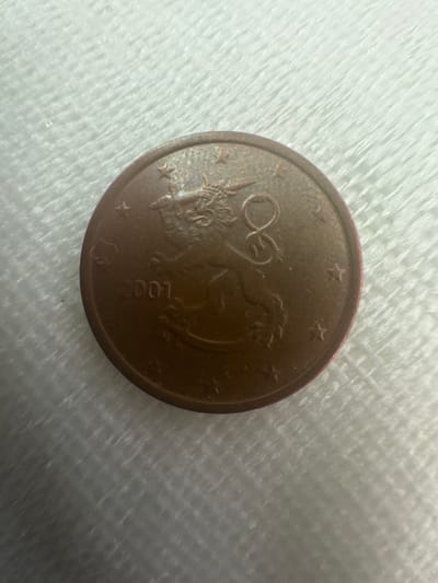 The image shows a single 5 euro cent coin from Finland, minted in 2001. The coin features the coat of arms of Finland, a crowned lion, and the denomination "5" encircled by twelve stars, which are a symbol of the European Union. The coin is made of copper-covered steel and has a diameter of 21.25 mm. The coin is placed on a white, textured surface, possibly a fabric or carpet. The background is out of focus, suggesting a shallow depth of field. There is no information in the image that indicates the location in the city of Schkopau, Germany.