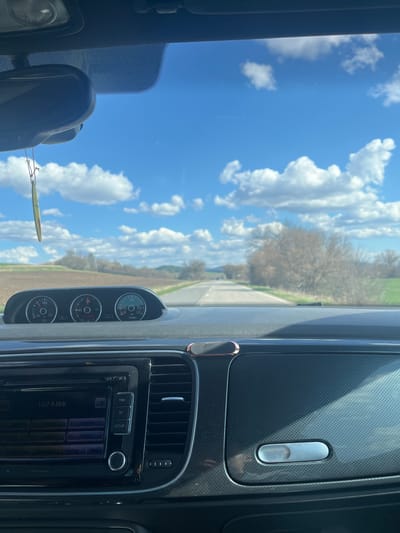The image shows the interior of a car driving on a road, with the dashboard, gauges, radio, and part of the windshield visible. Outside the car, there is a road, fields, trees without leaves, a blue sky, and fluffy white clouds. The radio frequency is 107.8 MHz. The gauges appear to measure oil temperature and turbo pressure. There is no information in the image that allows to determine the location as the city of Breznik, Bulgaria.