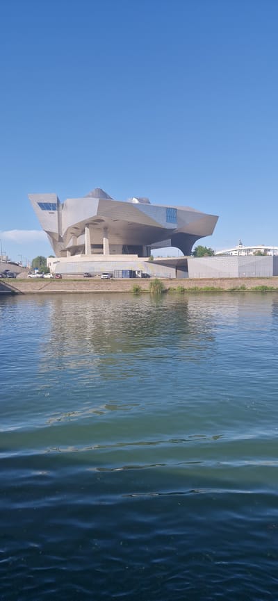 The image displays the Musée des Confluences, a modern architectural structure with a complex, faceted metallic exterior, situated on the bank of a body of water, likely a river, in the vicinity of La Mulatière, France. The building occupies the upper-middle portion of the frame, presenting multiple angles and reflective surfaces under a clear, bright blue sky. A subtle, wispy cloud is visible in the upper-left quadrant of the sky.
The museum rests on a raised foundation with visible support columns. Below the building, a concrete embankment runs parallel to the water. A road is present on this embankment, featuring several parked vehicles, including light-colored cars, a white van, and a blue storage container or shed. A small clump of green reeds or vegetation grows on the embankment to the right of the building. In the background on the far right, beyond the museum, additional distant buildings are discernible, one of which exhibits a distinct white, pointed roof structure.
The lower two-fifths of the image comprises a body of water with numerous ripples and reflections of the sky and the building's exterior. The water transitions in color from darker blues in the foreground to lighter blues and greens further back. No individuals are visible in the scene. The lighting conditions suggest a clear, sunny day.