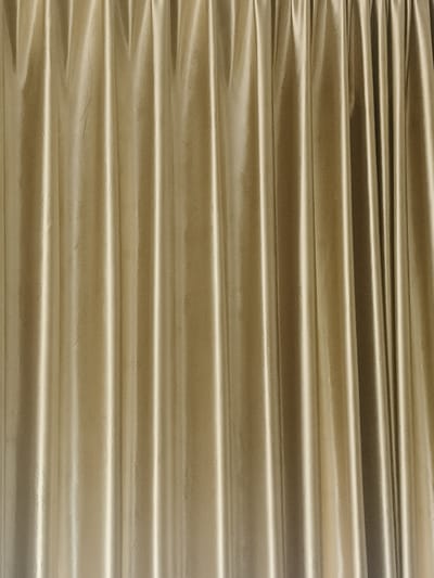 The media file shows a close-up, full-frame view of a golden-colored fabric, likely curtains. The fabric is heavily pleated, creating a textured surface with vertical lines and shadows. The lighting appears to be coming from the front and slightly to the left, highlighting the folds and giving the material a lustrous sheen. There are no discernible subjects, activities, or any text visible. The setting is abstract, focusing solely on the fabric's texture and color. Given the context of Chiang Rai City Municipality, Thailand, this could be a decorative element in a hotel, theater, or event venue. The time of day and weather are not ascertainable from the image.