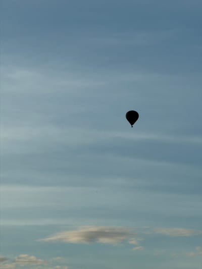 A single, dark silhouette of a hot air balloon is suspended in a vast, pale blue sky. The sky is streaked with faint, wispy clouds, suggesting either early morning or late afternoon light. The balloon appears to be centrally located in the frame, though slightly to the right. No specific landmarks or context of Naka, Laos are visible in this image, as it focuses solely on the balloon against the sky.