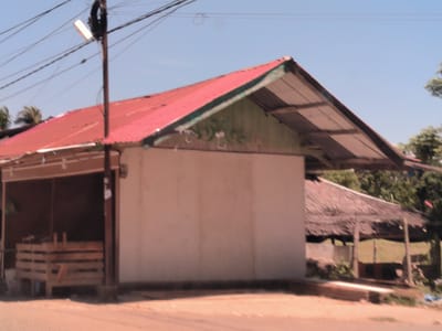 The image depicts a building with a red metal roof in Keudepanteubreuh, Indonesia. The building has a partially open side with wooden crates visible inside. Adjacent to the building is a structure with a thatched roof. A utility pole with wires is present in the foreground. The sky is light blue. The image quality is slightly blurry.