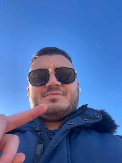 A male individual is depicted in a close-up, upward-angled outdoor shot, presumed to be a selfie, against a clear blue sky in Zarautz, Spain. He wears dark-lensed sunglasses with gold-toned frames and a dark blue winter jacket featuring a zipper, snaps, and a dark fur-trimmed hood. A white earbud is visible in his right ear. His left index finger is extended and pointed towards the lower-left foreground of the image. The subject has dark hair and a beard, presenting a neutral to slightly smiling facial expression. No other individuals or environmental details beyond the sky are visible.