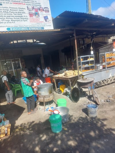 The image depicts a street-level view of a bustling outdoor market or shop in Tanzania, with a focus on traditional remedies. Several people are present, some appearing to be customers or vendors, with various objects related to food preparation and commerce scattered around. In the foreground, a banner with text in Swahili advertises "CHIKUNTILA," a natural remedy for a wide range of ailments. The banner also provides contact numbers and mentions availability in Picha Ya Ndege and Kariakoo.

Behind the banner, a stall or shop displays food items, possibly fried snacks, in glass cases. Various large metal pots, buckets, and containers are visible, suggesting cooking or storage activities. Some people are seated or standing near the stall, one individual is tending to a large metal drum, and another is looking at a tablet or phone. The ground is dusty, and shadows indicate it is likely daytime. The overall scene suggests a vibrant and active commercial environment where traditional health solutions are being offered.