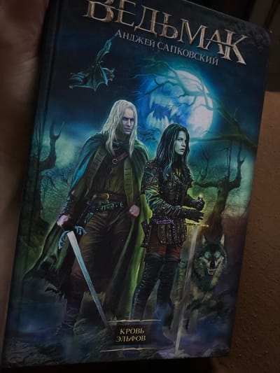 The image displays a physical copy of a book, likely a novel, featuring a dramatic fantasy illustration on its cover. The book is titled "ВЕДЬМАК" (The Witcher), written by "АНДЖЕЙ САПКОВСКИЙ" (Andrzej Sapkowski), with a specific volume title "КРОВЬ ЭЛЬФОВ" (Blood of Elves) visible vertically on the right side.

The cover art depicts a dark, mystical forest scene at night, dominated by a large, luminous full moon in the background. Two central figures, characteristic of fantasy heroes, are prominent. On the right, a woman with long dark hair, dressed in dark, elaborate leather armor and gauntlets, holds a sword. Her expression is serious and focused. To her left, a man with long white or very light blonde hair, clad in a dark green cloak over armor, also holds a sword. Both appear ready for combat or vigil.

In the upper right background, a snarling wolf with piercing, glowing green eyes emerges from the shadows, adding a menacing element. On the left side, closer to the book's spine, a small, dark, bat-like creature is visible in flight. The forest itself is depicted with bare, gnarled trees against a deep blue and green nocturnal sky, suggesting an ancient and possibly dangerous setting. The overall mood is one of dark fantasy, adventure, and potential conflict.

The book itself is slightly angled, resting on an indistinct, textured, dark brown surface, possibly fabric. The lighting is somewhat dim but adequately illuminates the cover's details and vibrant colors. The location context provided (Verkhnevesyoloye, Russia) suggests where this photograph was taken.