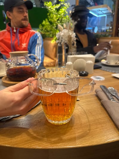 An indoor scene, likely a cafe or restaurant in Ul'yanovka, Russia, shows people gathered around a wooden table. The image is taken from a first-person perspective, focusing on a person's hand holding a unique glass mug filled with a clear, amber-colored beverage, possibly tea or juice. The mug features a distinctive lid with a hinged handle mechanism.
To the left, a man is visible wearing a red and blue Adidas track jacket with the brand's logo clearly displayed on his chest, and a black baseball cap. He appears to be looking towards the camera or another person at the table. Next to him, a clear glass teapot, also containing a dark liquid, rests on the table. In the background, further left, another person in dark clothing is partially visible, seemingly engaged with a phone or menu.
The table, made of light-colored wood with visible grain, is set with cutlery wrapped in a light napkin, white ceramic cups, and saucers. Decorative elements such as green foliage and dried ornamental plants are also present on the table. The lighting is warm and artificial, suggesting an evening or indoor setting. Reflections on the table surface indicate a glossy finish or slight dampness. The overall atmosphere appears casual and relaxed, with individuals enjoying drinks.