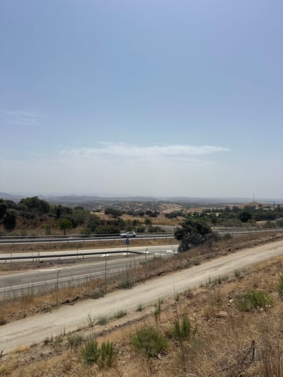 The image depicts a wide, hazy landscape with a bright, clear sky occupying the upper portion. In the middle ground, a highway runs horizontally across the frame, with a white car visible driving on it. Adjacent to the highway is a dirt road running parallel and slightly closer to the foreground. The foreground consists of dry, arid terrain with sparse, light-colored vegetation and soil. The background features rolling hills covered with scattered trees and vegetation, extending to a distant, hazy horizon. The scene suggests a rural or semi-rural location under a bright, possibly hot, day. No specific landmarks or identifiable structures indicative of Santa Elena, Spain are visible within the frame, making it impossible to confirm the location based solely on the image.