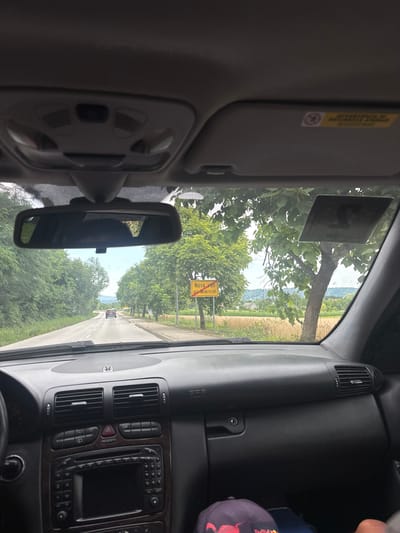The image is a first-person perspective from inside a car, looking forward through the windshield. The car is driving on a paved road bordered by lush green trees. In the distance, a yellow road sign reads "Nova vas pri Mokricah". The interior of the car is visible, including the dashboard with air vents and a car stereo. The sky is overcast, suggesting a cloudy day. There are no people clearly visible within the car. The surrounding environment appears to be rural or semi-rural, with agricultural fields visible beyond the trees.