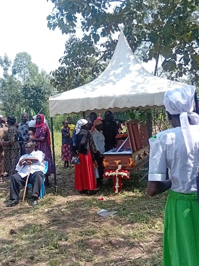 The image displays an outdoor gathering in a grassy area with trees in the background, located in Butali Chegulo ward, Kenya, under sunny conditions. A white peaked canopy tent is positioned centrally. Beneath the tent, an open wooden coffin with decorative handles rests on a stand. Adjacent to the coffin, a cross-shaped floral arrangement of red and white flowers lies on the ground.

Approximately twenty individuals are visible. In the mid-ground left, an elderly male subject sits in a blue plastic chair, dressed in a white short-sleeved shirt, dark trousers, and dark shoes, holding a wooden walking stick. He faces partially away from the coffin. Numerous individuals, including adults and some children, are gathered around the tent and coffin. One female subject in the mid-ground stands near the coffin, wearing a black top and a long red skirt. In the foreground right, the back of a subject is visible, wearing a white head covering, a white short-sleeved shirt, and a long green skirt. The ground consists of dry grass with patches of bare earth, and a piece of white debris, possibly paper or plastic, is visible near the floral cross.