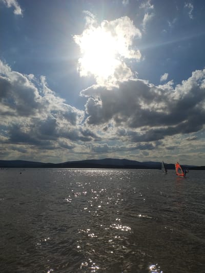 This is a vertically oriented, full shot image taken at Radslav, Czechia. The scene depicts a body of water with the sun shining brightly through dramatic clouds. The sun, a dominant feature in the upper center of the frame, casts brilliant rays and creates a shimmering effect on the water's surface. The water is dark and choppy, reflecting the sunlight in numerous small, bright patches. In the distance, a dark, silhouetted treeline or mountain range forms the horizon. On the water, there are several individuals engaged in watersports. Two windsurfers are visible: one with a white sail and another with a bright orange sail. Other figures can be seen further in the distance on the water, possibly paddleboarding or swimming, indicated by small dark shapes. The sky is a mix of bright blue and heavy, dark gray clouds, creating a dynamic and slightly ominous atmosphere despite the bright sunshine.