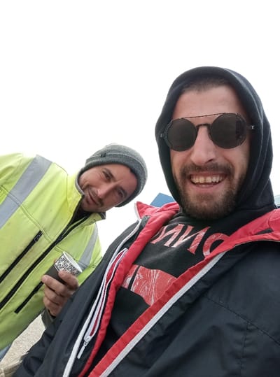 This image captures two adult males outdoors in Drugan, Bulgaria, engaged in a selfie-style photograph. The foreground subject, positioned closer to the camera and likely holding it, is a male with a beard and moustache, wearing a black hooded sweatshirt with the hood up, dark round sunglasses, and a dark outer jacket with red lining and white zippered accents. He exhibits a broad smile, showing teeth, and looks directly at the camera. The subject behind and to the left is an adult male with short dark hair, a slight beard and moustache, wearing a grey knit beanie and a bright neon yellow high-visibility jacket with grey reflective strips on the shoulders and arms, featuring black zippers. He holds a dark-colored disposable cup with a white intricate pattern in his right hand and has a subtle smile while looking towards the camera. The background is dominated by an overexposed white sky, indicative of an outdoor setting on an overcast day, with a small portion of a blue structure visible behind the foreground subject. The ground in the lower left appears to be a light-colored paved surface.
