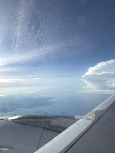 The image is a photograph taken from an airplane window, looking out at the sky and the ground below. The sky is a bright blue with scattered clouds. The ground appears to be a landscape of islands and water, possibly the coast of Thailand. The airplane wing is visible in the foreground, and the window has some smudges and streaks on it. The image is taken from a high altitude, and the light is bright, suggesting it is daytime. The location is Sisa Chorakhe Noi Subdistrict, Thailand.