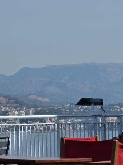 The image shows a distant view of the city of Magaluf, Spain, with a hazy mountain range in the background. In the foreground, a white metal railing separates the viewer from the water. Several red chairs are positioned behind the railing, suggesting an outdoor seating area, likely on a ferry or a waterfront establishment. A black outdoor lamp is mounted on a pole just behind the railing. The water is calm and reflects the hazy sky. Buildings of various sizes and colors are visible along the coastline of Magaluf, nestled between the water and the hills. The overall impression is one of a sunny day with a somewhat muted visibility due to atmospheric conditions.