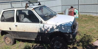The image shows a white, four-door hatchback vehicle parked outdoors. The car is old and appears to be in disrepair, with visible rust on the body and a damaged front bumper. It has a roof rack with what appears to be a light bar attached. The car is parked on a grassy area with some dirt patches, next to a grey metal fence. In the background, there are some outbuildings, including a small, red and green structure that resembles a dog house or a small shed, and a metal bin. The scene is illuminated by sunlight, casting shadows from trees onto the car and the ground. The location is specified as Esipovo, Russia.