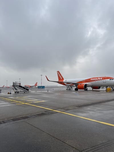 The scene depicts an airport tarmac at Milan Malpensa Airport in Somma Lombardo, Italy, under an overcast, uniformly gray sky. The asphalt surface is wet with standing water and reflections, consistent with recent precipitation.
In the mid-ground, a white and orange EasyJet Airbus A320 series aircraft is parked, positioned diagonally with its nose facing towards the left. Passenger boarding stairs are deployed and connected to both the forward and aft main passenger doors. Two individuals wearing yellow high-visibility clothing are visible on the tarmac near the aircraft's front landing gear.
To the left foreground, a standalone silver mobile passenger stair unit, featuring red and white checkered safety markings on its top platform, rests on the wet tarmac. Multiple other EasyJet aircraft in identical livery are visible parked in the distance, indicating an active airport apron. Yellow operational markings are painted on the tarmac surface, defining various areas. Tall utility poles are scattered across the apron.