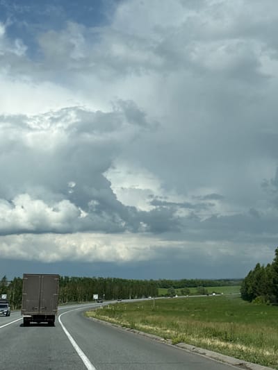 The image depicts a highway scene in a rural area, potentially near Kendektamak, Russia, characterized by a gray, overcast sky with dramatic clouds. A dark brown truck is in the foreground, traveling on a paved road that curves to the right. The road has white lane markings. To the right of the road is a grassy field with some scattered trees and a dense line of trees in the background. Several other vehicles are visible on the highway in the distance, including cars and possibly other trucks. The overall atmosphere is somber due to the cloudy sky, suggesting impending rain or a recent storm.