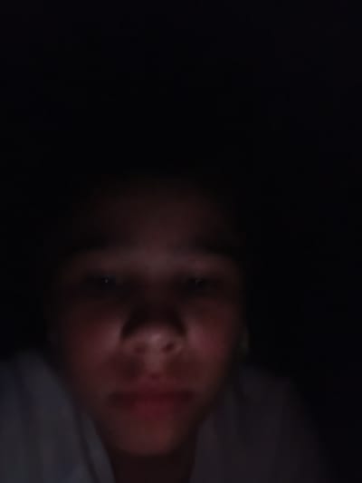 The image shows a close-up of a person's face in very low light conditions. The person's eyes, nose, and mouth are visible, with their face illuminated by a soft, reddish light source, possibly a screen. The background is predominantly dark. There are no discernible elements in the image that indicate a specific location within the city of Opoczno, Poland, or any other city. The image is a selfie or a similarly framed personal photograph, not a scene set within a particular urban environment.