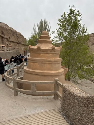 This image, rotated 90 degrees clockwise, depicts an outdoor cultural or historical site in Guazhou County, China, under an overcast sky. The central subject is a large, light-brown, multi-tiered architectural structure, resembling a decorative stupa or column, characterized by its distinct curved and stacked segments.

In the midground, a group of people, appearing to be tourists or visitors, are seen walking along a concrete pathway and ascending a series of steps. They are dressed in casual attire, suggesting a leisurely exploration of the site. A robust concrete railing with a textured, pebbled top surface runs alongside the path, serving as a safety barrier.

The background reveals a dramatic, rugged cliff face, into which numerous wooden walkways, stairs, and structures are integrated, hinting at ancient cave dwellings or temples carved into the rock. Patches of vibrant green foliage from trees with visible branches are interspersed across the landscape, providing a natural contrast to the muted tones of the rock and sky. The sky itself is a soft, uniform gray, indicating an overcast day with diffused lighting. The overall atmosphere is calm, with visitors experiencing the historical environment. There is no visible text in the image.