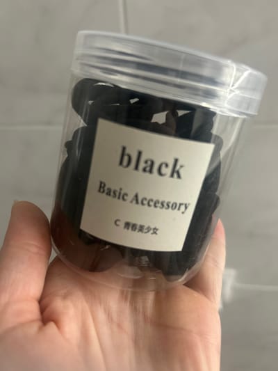 A clear plastic container filled with black hair ties is held by a person's hand. A label on the container reads "black Basic Accessory" in English, with Chinese characters below. The background appears to be a tiled wall. The image does not contain any information about the city of Нижние Муллы, Russia.