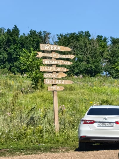 The image depicts a directional signpost in the city of Naumovka, Russia. The signpost is made of wood and has several arrow-shaped planks pointing in various directions. These planks are inscribed with Russian text, indicating points of interest. The text reads "ПАСЕКА" (Apiary), "КАТАНИЕ НА ЛОШАДЯХ" (Horseback Riding), "СТРЕЛЬБА ИЗ ЛУКА" (Archery), "КВАДРОЦИКЛЫ" (ATVs), "КУМЫС" (Koumiss), "БЕСЕДКИ" (Gazebos), and "МЕД" (Honey). The signpost stands in a grassy, field-like environment with trees in the background and a clear blue sky above. A white car is partially visible in the lower right corner of the image, its rear license plate reading "M580BM 702". The scene appears to be an outdoor recreational area.