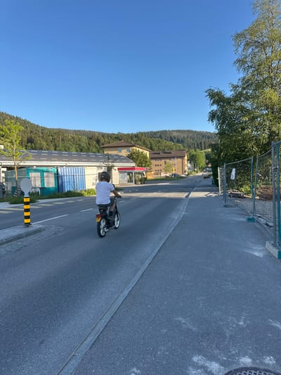 A person wearing a white t-shirt and shorts, a helmet, and dark shoes rides a moped on a paved road in Le Brassus, Switzerland. The moped has a yellow license plate with red lettering. The person is viewed from behind, and they are riding towards the right side of the frame. The road is lined with a sidewalk on the left, which has a yellow and black striped pole with a white circular sign. On the right side of the road, there is a construction fence with some wooden pallets behind it. The background features buildings with solar panels on their roofs, trees, and a forested hill under a clear blue sky.