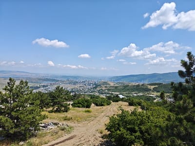 This is a high-angle, wide shot of a landscape with a city in the distance. The foreground is a dry, dirt path with some scattered debris, including pieces of wood and metal. Green pine trees are prominent on the left and right sides of the frame, with dry grass and some smaller green bushes in between.
In the midground, rolling hills covered in greenery and sparse trees descend towards a city. The city is densely packed with buildings of varying heights, including some taller, modern structures. A body of water, possibly a lake or reservoir, is visible within the city.
The background features more distant, hazy hills and mountains under a bright blue sky with scattered white cumulus clouds. The overall impression is of a clear, sunny day, likely in the summer or early autumn, with a view overlooking a city from a natural vantage point. The scene is devoid of any people or animals. The location is identified as Mukhatskaro, Georgia.