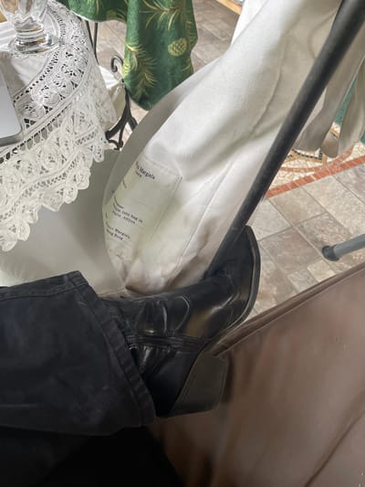 The scene shows a black leather boot, partially obscured by black pants, resting on a white cotton Maison Margiela tote bag. The tote bag has a tag with printed text. Nearby is a white lace object. The background includes a brown surface and tiled flooring.