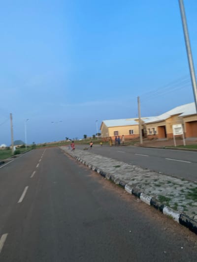 The image depicts a street scene in Dumnga, Nigeria, likely captured during the early morning or late afternoon due to the soft, blue sky and long shadows. The perspective is from a low angle, looking up and to the side, with the road taking up a significant portion of the frame and tilting diagonally.
On the left side of the road, there are buildings with light-colored facades, appearing to be residential or commercial structures. Some have visible windows and architectural details. A light pole with wires runs along the side of the road, suggesting it's a developed area. The road itself is asphalt with white dashed lines indicating lanes.
The right side of the road features a gravelly area with some greenery, bordered by a curb with alternating black and white markings. Further down the road, a few individuals can be seen walking or standing, their activities not clearly discernible due to the distance and angle. One person appears to be wearing a red garment.
The overall impression is of a quiet, everyday scene in a Nigerian town. The weather appears clear and calm. There is no visible text in the image.