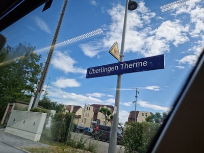 The image, taken from inside a vehicle looking outwards through a window, shows a street scene in Überlingen, Germany. A prominent blue sign with white text reads "Überlingen Therme," indicating the location. To the left of the sign, a yellow triangular warning sign with an illustration of a train is visible. The sky is blue with scattered white clouds. In the background, several buildings with reddish-brown roofs are visible, some appearing to be houses or apartments. A street is present in the foreground, with parked cars including a black van and a blue car. A chain-link fence separates the street from a grassy area with some bushes and trees. A tall pole with a light fixture and some electrical equipment is also visible. The scene appears to be captured on a sunny day.