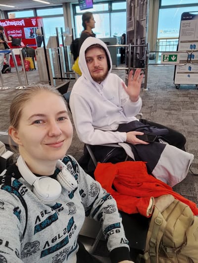 The image depicts two individuals seated in what appears to be an airport departure lounge in Yagel, Israel.
In the foreground, a light-skinned female with light brown hair is positioned towards the left, looking directly at the camera with a slight smile. She is wearing a grey long-sleeved sweater with teal and black patterns and has white over-ear headphones draped around her neck. Her left hand is visible, resting on her lap.
To her right and slightly behind, a light-skinned male with a beard and light brown hair is seated, looking towards the camera and raising his right hand in a waving gesture. He is wearing a white hooded sweatshirt with the hood up, and dark trousers. His left hand holds a dark-colored smartphone. A dark blue and white jacket is draped over his lap.
Adjacent to the female, on an empty seat, a folded red garment and a tan/khaki backpack are present. The floor is covered with a grey patterned carpet.
In the background, beyond metal queue barriers, large windows offer a view of an outdoor area, possibly an airport tarmac. Several other individuals are visible in the background; one person in a dark jacket walks near a red information kiosk with text partially visible as "Passport Ready" and "Don't Give Up Your Dreams". Another person with dark hair walks further back near the windows. A prominent white sign with black Hebrew text, including "מידע לנוסע" (Information for the Traveler) and an emergency exit symbol, is visible on the right side of the background, confirming the Israeli location.