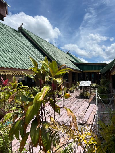 The image shows an outdoor wooden deck area with several potted plants in the foreground and middle ground. The deck is made of weathered wooden planks. In the background, there is a building with a green corrugated metal roof and decorative wooden trim. The sky is blue with scattered white clouds. The scene appears to be a serene outdoor seating or walkway area. The image is a clear, eye-level shot, with no people visible. The location is Tambon Ko Panyi, Thailand.