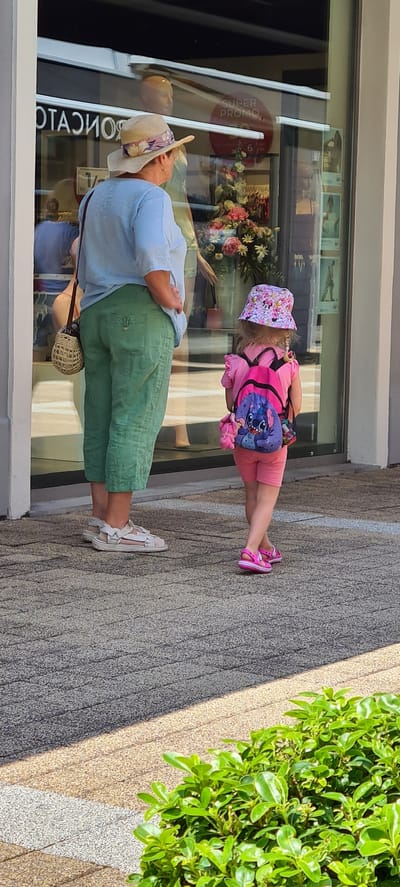 The image shows two people, likely a child and an adult, standing on a textured pavement in front of a shop window. The adult, wearing a light blue shirt, green pants, and a wide-brimmed straw hat decorated with floral fabric, is seen from the back and side. They are carrying a small woven bag. The child, facing away from the camera, is dressed in a pink outfit including a t-shirt, shorts, and pink sandals. They are wearing a pink bucket hat with Mickey Mouse characters and a backpack featuring the character Stitch. The child's legs are bent as if in mid-stride.
Reflected in the shop window are displays of flowers and mannequins, suggesting the shop sells clothing or accessories. A red circular sticker with white text is also visible on the window, reading "SUPER PROMO". The lighting suggests it is daytime, with shadows cast on the pavement. The overall scene appears to be outdoors, possibly on a sunny day, in a pedestrian area. The individuals' clothing and accessories indicate a casual, possibly warm weather outing.