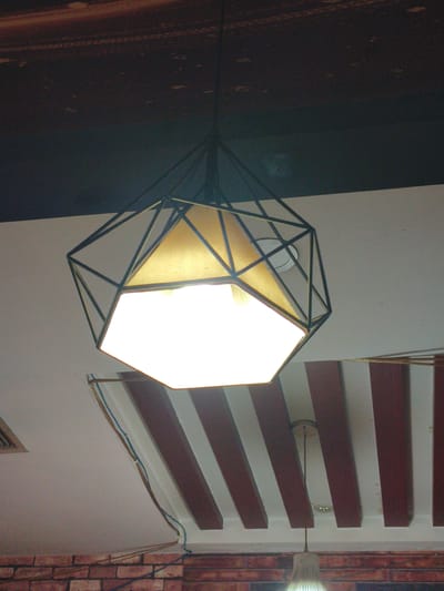 A modern geometric pendant light with a black metal frame and a hexagonal shade hangs from a dark ceiling. Below the light, the ceiling is white with a decorative element featuring alternating dark red and white stripes. Wires run along the white ceiling, attached by metal fixtures. The background shows a hint of a brick wall and another light fixture. The scene appears to be indoors, likely in a commercial space, with no people visible. The location of Jixi, China, is not discernible from the image itself.