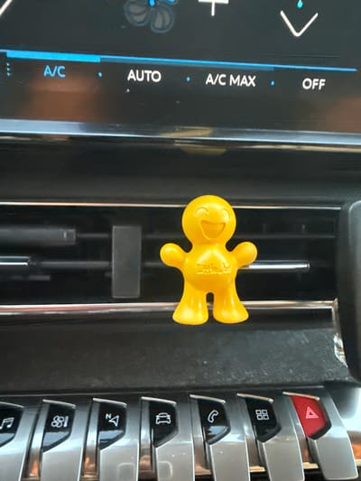 The image shows a yellow plastic air freshener shaped like a little person clipped to a car's air vent. The air vent is part of the dashboard of a car. On the dashboard, there are digital controls for the car's climate system, displaying options like "A/C", "AUTO", "A/C MAX", and "OFF". Below the air vent, there are several physical buttons with icons, including a music note, a phone, a grid of squares, and a hazard light symbol. The lighting suggests the photo was taken inside the car during the daytime. The image does not contain any information related to Longpont-sur-Orge, France.