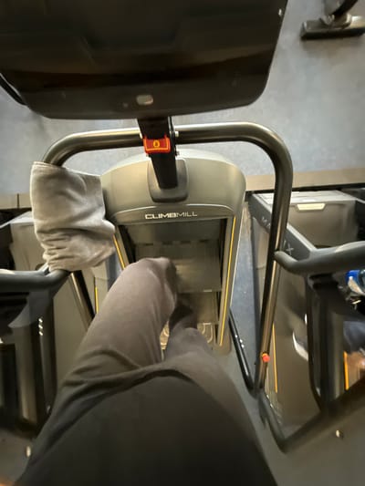 In Temse, Belgium, the image depicts a first-person perspective of an individual exercising on a "CLIMBMILL" stair-climbing machine within what appears to be a fitness center. The primary subject is the lower torso and legs of a person, clothed in dark gray or black athletic pants, actively stepping onto the machine's moving steps.
The "CLIMBMILL" machine, visible from the console downwards, is predominantly dark gray with yellow accent lines on the sides of its steps. A light-colored, possibly grey, towel is draped over the left handle. A red and orange safety clip, marked with a white "0", is attached to a vertical support pole below the main console, which is partially visible at the top of the frame. The machine's dark, metallic handles curve upwards on both sides.
To the right of the "CLIMBMILL," another piece of exercise equipment, identified by the partial brand name "MATRIX," is visible. This machine also features dark framing. The floor is a uniform dark gray, and in the upper right background, a vacuum cleaner head with a silver handle is resting against a dark wall or surface, suggesting a cleaning presence in the facility. No other individuals or direct interactions are visible. The environment appears utilitarian and consistent with an indoor gym setting.