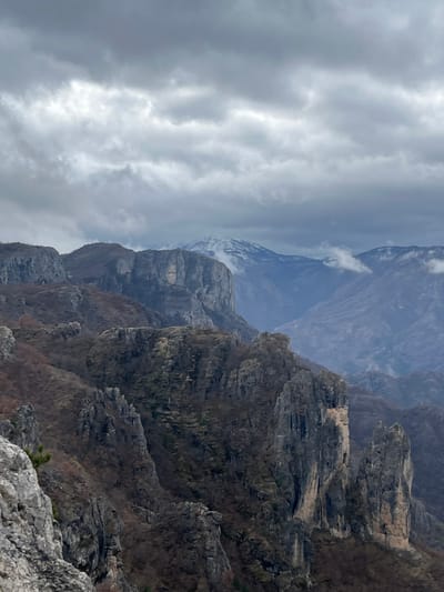 The scene depicts a panoramic view of a rugged, mountainous landscape in Klobuk, Bosnia and Herzegovina, under an overcast sky. In the immediate foreground, a light grey, jagged rock formation with a solitary pine branch is visible at the lower left. The midground features multiple layers of imposing, dark grey and brown rock cliffs and peaks, characterized by vertical faces, deep crevices, and a sparse covering of dormant, reddish-brown vegetation. Prominent, slender rock pinnacles rise from the terrain in the lower right and center. In the background, successive ranges of mountains recede into the distance, appearing progressively bluer due to atmospheric perspective. The furthest mountains to the left exhibit visible patches of snow on their peaks, while wispy clouds partially obscure some distant slopes. The sky above is entirely covered by a dense layer of dark grey stratus clouds. There are no individuals, man-made structures, or observable actions within the frame.