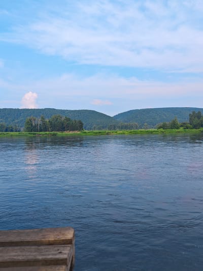 A calm river flows in front of a lush, tree-covered hillside under a cloudy sky. In the foreground, the corner of a wooden structure, possibly a dock, juts into the water. The water is dark blue with ripples reflecting the sky and surrounding landscape. The hillside is covered in dense green trees, with a lighter patch of trees on a small island in the middle distance. The sky is a soft blue with scattered white clouds. The overall scene depicts a peaceful, natural landscape. The location is the city of Yukseevo, Russia.