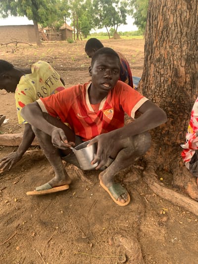 A young Black male is squatting on the ground next to a large tree. He is wearing a red and orange striped t-shirt and a single sandal on his left foot. He is holding a small metal bowl in his right hand and a spoon in his left hand. He appears to be eating from the bowl. Other people are visible in the background. The ground is dirt and there are some scattered leaves and twigs. In the background, there are some simple mud-brick structures and trees. The overall setting appears to be rural, possibly a village in Dull A, Nigeria.