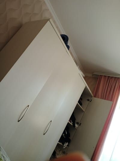 This image depicts the interior of a white wardrobe with its right door ajar. The wardrobe is filled with various items, including clothing. One shelf is visible, containing folded dark garments and a pair of white shoes. The wardrobe is situated in a room with a textured beige wallpaper on the wall and a white ceiling. To the right of the wardrobe, a pink curtain is visible, partially obscuring a bright window. In the upper left corner, a dark blue rolled-up object, possibly a yoga mat, rests on top of the wardrobe. The image does not provide any specific context to pinpoint its location in Kuntuu, Kyrgyzstan, but it appears to be a domestic setting.