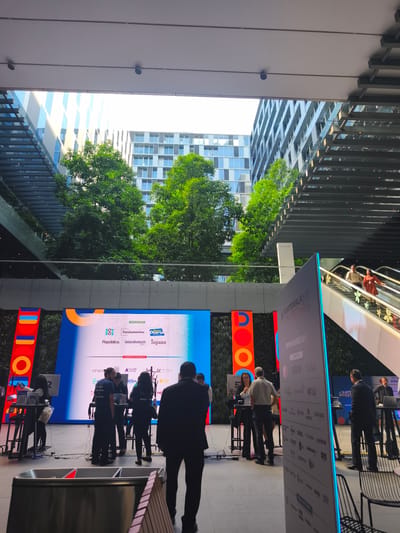 The image shows an event occurring outdoors in Guatemala City, Guatemala, featuring a stage with a large screen displaying logos and event sponsors, including "SOY 502," "Fundamentos," and "latamfintech HUB." People are gathered around the stage, with some standing at tables and others engaged in conversation. There is a sponsored poster to the right with the text "GUATEMALA >20 FINTTECH SHOT>26" visible on it. An escalator is visible in the background, and trees are between the buildings. The location appears to be a modern building complex with open areas.