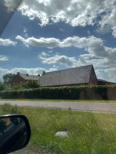 The image shows a view of a rural landscape in Wierden, Netherlands, under a partly cloudy sky. In the foreground, a car's side mirror reflects a person wearing a blue shirt. Beyond the mirror, tall green grass lines a paved road. Behind the road, a dense, well-maintained hedge forms a barrier in front of a long, red-brick building with a grey tiled roof. The building appears to be a farmhouse or a similar rural structure, with visible chimneys and windows. Trees are interspersed with the building and behind the hedge, suggesting a lush environment. The overall scene conveys a sense of a peaceful day in the Dutch countryside.