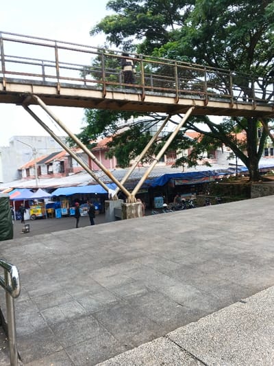 This image depicts an outdoor urban scene in Sarandi de Cuadra, Uruguay, characterized by a multi-level layout featuring a pedestrian bridge, a bustling market area below, and residential/commercial structures in the background, all under a bright, overcast sky.
In the foreground, a gray concrete tiled surface serves as a viewing platform, with a silver metal handrail partially visible on the bottom left, indicating a stairway or ramp descent. Dominating the midground is an elevated pedestrian bridge, constructed from aged metal with a flat deck and supported by multiple V-shaped metal beams resting on concrete bases. A single individual, clad in a horizontally striped top and dark pants, is observed walking from left to right on the bridge.
Below the bridge and the viewing platform lies an active market area. Multiple temporary stalls are present, distinguished by various colored tarpaulin roofs, predominantly blue, with some red and white. Visible market objects include a yellow food cart on the far left and several blue vending kiosks. Two individuals are central to this area, standing and appearing to interact; one wears a red cap. Further right, a group of motorcycles or scooters is parked.
The background reveals a mix of buildings with visible red-tiled roofs and lighter-colored walls, interspersed with lush, large green trees, particularly a prominent tree on the right side of the frame whose canopy extends across the upper right quadrant, intertwining with the bridge structure. The overall environment suggests a lively public space.