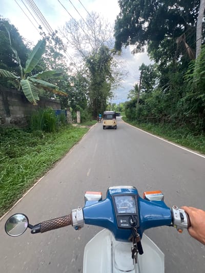 A point-of-view shot from a blue and white Honda motorcycle on a paved road outside Angulugaha Junction, Sri Lanka. A beige tuk-tuk is visible ahead in the distance. The foreground includes the motorcycle's handlebars and a partial view of the driver's right hand. Lush green vegetation borders the roadside. Overhead, trees and power lines are visible against a cloudy sky. A banana plant is visible along the lower left edge.