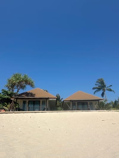 The scene depicts a sunny daytime view of a sandy beach in Phra Ae, Thailand, with two identical bungalows positioned on elevated ground beyond the sand. The foreground features light beige sand, lightly scattered with small white debris. Above the sand, two single-story bungalows with light brown tiled, pitched roofs and white exterior walls are visible. Each bungalow has a front balcony with dark-framed sliding glass doors and a railing. A multi-trunked tree with spiky, elongated green leaves stands to the left of the left bungalow. Various green foliage is present behind and between the bungalows. To the right of the right bungalow, a tall palm tree with green fronds is visible. The sky above is clear and uniformly bright blue. No people, discernible actions, or interactions are visible in the image.