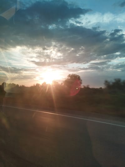 This image depicts a sunset scene viewed from a moving vehicle on a road. The sun is low in the sky, casting a bright glare and creating lens flare effects. Dark clouds are present in the upper portion of the sky, contrasting with the bright sunlight breaking through. The foreground shows a road with a white line marking the lane. The middle ground features a silhouette of trees and vegetation, with a noticeable red lens flare on a tree. The overall impression is one of a journey during the golden hour, likely near Ahunbabaev, Uzbekistan, given the context.