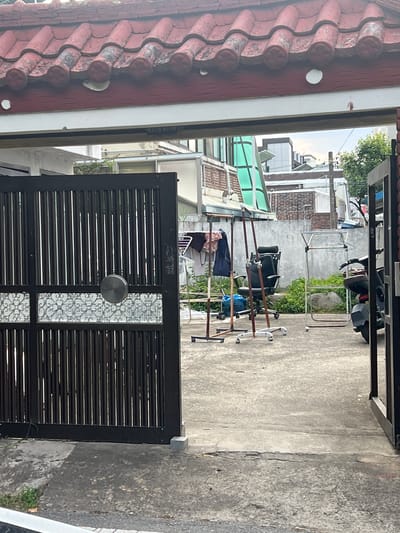 The image shows an open black metal gate with decorative latticework in the lower panel, leading into a concrete courtyard. To the left, a portion of a red tiled roof is visible. Inside the courtyard, there are several objects, including clothes racks with laundry hanging on them, an office chair on wheels, a blue plastic bag, and a motorcycle parked on the right. A grey concrete wall forms the background. The scene appears to be an outdoor area of a residential or commercial property in Gwangju, South Korea. The sky is overcast.